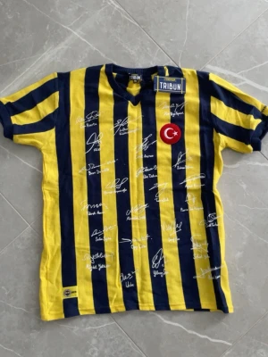 Fenerbahce matchtröja med autografer - Säljer en Fenerbahce matchtröja i gul och marinblå med vertikala ränder. Tröjan har autografer på framsidan och ett turkiskt flaggmärke på bröstet. Klassisk sportig look med korta ärmar och rund hals. Den är väldigt sällsynt.