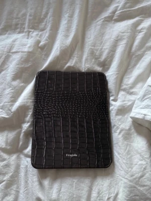Svart iPad-fodral från Fragola - Snyggt svart iPad-fodral från Fragola med krokodilpräglat mönster. Utsidan är i konstläder och insidan har en skyddande plastdel för att hålla din iPad på plats. Fodralet har öppningar för kamera och knappar. iPad A16 % iPad 10 gen