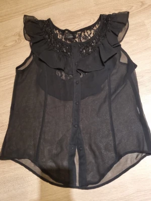Svart transparent blus med spets - Snygg svart ärmlös blus i transparent material med volang och spetsdetaljer vid halsringningen. Knäppning framtill och rundad nederkant. Perfekt att styla med ett linne under för en trendig look.