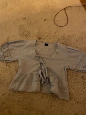 Ljusblå sommarblus med knyt och volanger - Supersöt ljusblå blus med knyt framtill och volangkant. Blusen har korta puffärmar med fina spetsdetaljer och rynkade avslut. Tillverkad i ett lätt och luftigt tyg, perfekt för en trendig och avslappnad look.