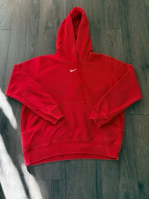 Röd vintage Nike hoodie med huva - Snygg röd hoodie från Nike med klassisk vit logga på bröstet. Hoodien har en stor ficka framtill, justerbar dragsko i huvan och ribbade muddar vid ärmar och nederkant. Perfekt för en avslappnad och sportig stil. Stolek XL men krympt något så passar nog M