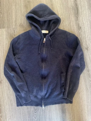 Mörkblå cashmere hoodie från Sayles - Mysig mörkblå hoodie i cashmere från Sayles med dragkedja framtill och två fickor. Klassisk huva med dragsko och ribbade muddar vid ärmslut och nederkant. Perfekt för dig som gillar stilren och bekväm streetwear. Storlek M men är liten i storleken så passar S