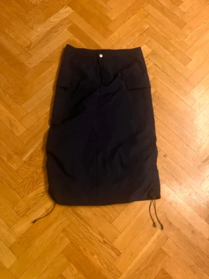 Mörkblå kjol med snörning från SHEIN - Mörkblå långkjol från SHEIN med snörning i nederkanten och knappdetalj i midjan. Kjolen har en enkel, rak passform och är tillverkad i ett lätt syntetmaterial. Perfekt för dig som gillar stilrena plagg med en twist.Skriv gärna för mått! 💕