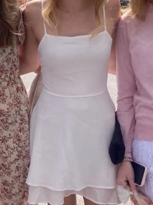 Vit kortklänning med snörning - Supersöt vit kortklänning med tunna axelband och öppen rygg med snörningsdetalj. Klänningen har en lätt utsvängd kjol och dubbla lager tyg för extra flow. Perfekt för sommarens alla tillfällen!