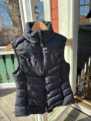 Svart dunväst med hög krage - Stilren svart dunväst med hög krage och dragkedja framtill. Västen har två sidofickor med dragkedja och quiltade partier för extra värme och en sportig look. Materialet är glansigt och lätt, perfekt att bära över en hoodie eller tröja under kyliga dagar.