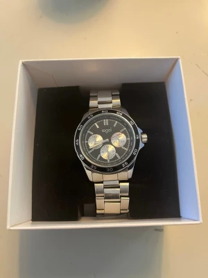 Silverfärgad klocka från Regal - Stilren klocka från Regal med silverfärgad länk i metall och svart urtavla. Klockan har tre dekorativa subdials och markerade siffror runt boetten. Perfekt accessoar för dig som gillar en klassisk och sportig look.