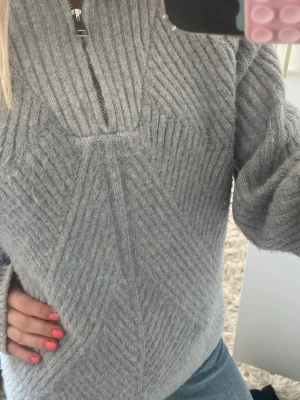 Ljusgrå stickad tröja med dragkedja - Mysig ljusgrå stickad tröja med halv dragkedja framtill och ribbade detaljer. Tröjan har ett snyggt mönster i stickningen och långa ärmar. Så nice till sommaren/vårkvällar 