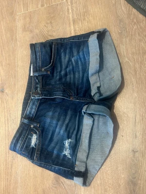 Mörkblå lågmidjade jeansshorts med upprullad kant  - Snygga mörkblå lågmidjade jeansshorts med upprullad kant och slitna detaljer framtill. Klassisk femficksmodell med knapp och dragkedja. Perfekta för varma dagar och enkel att matcha med olika stilar.