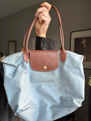 Ljusblå Longchamp Le Pliage väska - Säljer en ljusblå Longchamp Le Pliage handväska med bruna läderhandtag och lock. Väskan är tillverkad i slitstark nylon med detaljer i brunt läder och har en klassisk, rymlig form. Perfekt för dig som gillar stilrena och tidlösa accessoarer.