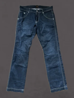 Mörkblå raka jeans med kontrastsömmar - Säljer ett par mörkblå jeans med raka ben och vita kontrastsömmar. Jeansen har fem fickor, silverfärgade knappar samt tydliga sömmar på både fram- och baksida. Materialet är klassisk denim och passformen är straight 