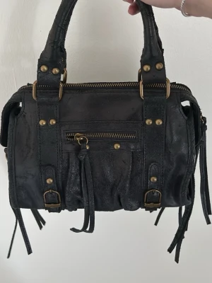 Svart handväska med nitar och detaljer - Cool svart handväska i läder med guldfärgade nitar och dragkedjor. Väskan har både handtag och avtagbar axelrem, samt dekorativa remmar och tofsar. Praktisk ytterficka med dragkedja framtill och snygga metalldetaljer.