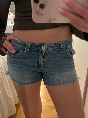 Blå denimshorts med low waist  - Säljer ett par blå denimshorts med rå, fransig kant och klassisk femficksdesign. Shortsen har låg midja, bälteshällor och knappgylf. Perfekta för varma dagar och ger en avslappnad look.