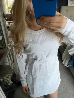 Vit långärmad klänning med bälte - Säljer en vit långärmad klänning från Aimin med rund hals och knytbälte i midjan. Klänningen har en enkel och clean design med logotyp på bröstet. Perfekt för dig som gillar sportiga och avslappnade outfits.