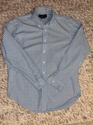 Blårutig skjorta från Ralph Lauren - Klassisk blårutig skjorta från Ralph Lauren i modellen Classic Fit. Skjortan har långa ärmar, knappar framtill och en tidlös krage. Tillverkad i mjuk bomull och passar perfekt till både jeans och chinos. Skjortan är large men något liten i modellen. Axelbredd:50 längd:80.