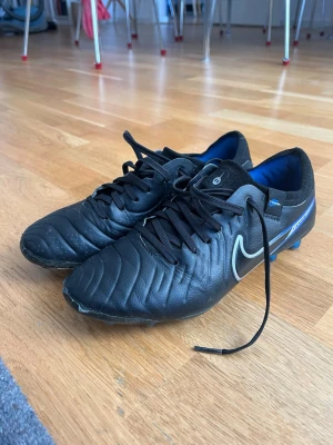 Nike tiempo PRO fotbollsskor för konstgräs - Nike tiempo fotbollssskor för konstgräs. Svarta och blåa AG fotbollsskor. Använda ungefär en halv säsong så han inga defekter som påverkar känslan. Bara några synliga. Storlek 37,5. Nypris 1000kr. 