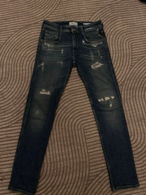 Replay anbass med slitningar - Säljer ett par blå skinny jeans från replay med slitningar och distressed detaljer framtill. Klassisk femficksmodell med snygga sömmar på bakfickorna och läderpatch i midjan. Perfekta för en avslappnad och trendig look.