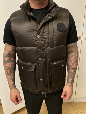 Mörkbrun dunväst från Snoot - Snygg mörkbrun dunväst från Snoot med hög krage, två stora fickor med lock och tryckknappar samt en bröstficka med dragkedja. Västen har ett stilrent märke på bröstet och är tillverkad i glansigt material. Perfekt för lager-på-lager-stil. Nypriset är 3500 kr och den är i perfekt skick. Näst intill ny 