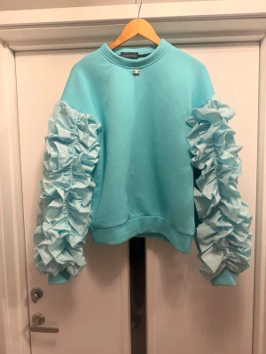 Turkos sweatshirt med volangärmar - Supersnygg turkos sweatshirt från Lavish Alice med unika volangärmar som verkligen sticker ut. Tröjan har rund halsringning och är gjord i mjukt material, perfekt för dig som vill ha något extra i garderoben. Volangerna på ärmarna ger en cool och trendig vibe. Från Polen!
