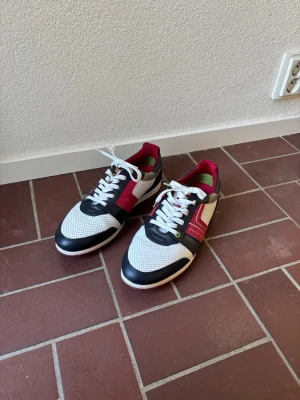 Vita och svarta sneakers från HUGO BOSS - Snygga sneakers från HUGO BOSS i vitt, svart och rött med gröna detaljer. Skorna har perforerat material på ovandelen, vita snören och en låg siluett. Märkeslogga syns på sidan. Perfekta för dig som gillar stilrena och sportiga skor.