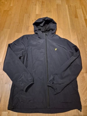 Svart vindjacka från Lyle & Scott - Snygg svart vindjacka från Lyle & Scott med huva och dragkedja framtill. Jackan har elastiska muddar vid ärmsluten och dragsko i midjan. Köpte den I Lyle & Scott butiken förra sommaren och är sparsamt använd. Nypriset var 1100 kr