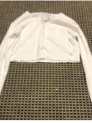 Vit cropped cardigan med knappar - Snygg vit cropped cardigan med långa ärmar och knäppning framtill. Enkel och stilren design som passar perfekt till lager på lager. Tillverkad i mjukt tyg för en bekväm känsla.