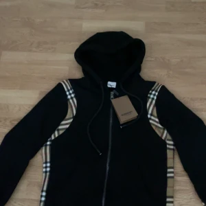 Svart Burberry kofta med rutiga detaljer - Snygg svart kofta från Burberry med dragkedja framtill och klassiska beige-rutiga paneler längs sidorna och ärmarna. Tröjan har huva med snörning och ribbade muddar. Perfekt för dig som gillar streetwear och vill ha något som sticker ut.