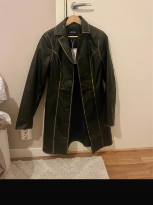 Svart lång skinnjacka från Monki - Stilren svart lång skinnjacka från Monki med markerade sömmar i kontrastfärg. Jackan har klassisk krage, knäppning framtill och en rak, något längre passform som ger en cool och trendig look.