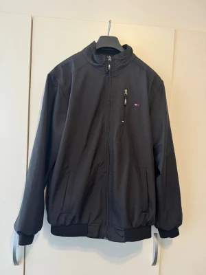 Svart vindjacka från Tommy Hilfiger - Snygg svart vindjacka från Tommy Hilfiger med hög krage, dragkedja framtill och en bröstficka med dragkedja. Jackan har diskret logga på bröstet och muddar vid ärmslut och nederkant. Tillverkad i lätt material som passar perfekt till vardagslooken.