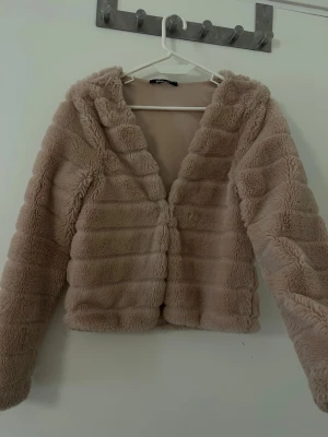 Beige fluffig pälsjacka - Mysig beige pälsjacka med horisontella sömmar och fluffig struktur. Jackan har en öppen front utan synliga knappar och en mjuk insida. Perfekt för kalla vinterdagar och ger en trendig look.