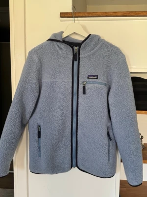 Ljusblå fleecehoodie från Patagonia - Mysig ljusblå fleecehoodie från Patagonia med dragkedja framtill, huva och tre fickor med dragkedja. Tillverkad i fluffigt, värmande material och har mörkblå detaljer vid dragkedjor och kanter. Perfekt för kalla dagar och outdoor-häng. I storlek S passar en större XS och S i helt!