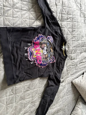 Svart Kenzo sweatshirt med tigertryck - Cool svart sweatshirt från Kenzo med ikoniskt tigerbroderi i färgerna rosa, gult, blått och vitt på bröstet. Rund halsringning med ribbade muddar och Kenzo Paris-tryck. Perfekt statement-plagg för dig som gillar streetwear och snygga detaljer.