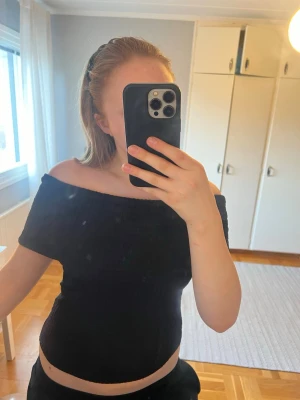 Svart offshoulder topp - Trendig svart offshoulder topp med smockad struktur och tight passform. Toppen är ärmlös och har en bred kant som sitter snyggt över axlarna. Perfekt att styla med både jeans och kjol för en cool look.