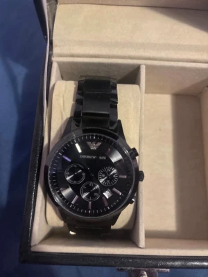 Svart klocka från Emporio Armani - Stilren svart klocka från Emporio Armani med metallänk och rund urtavla. Klockan har flera små urtavlor och tydliga markeringar, samt Emporio Armani-loggan högst upp. Perfekt accessoar för dig som gillar en klassisk men modern look.