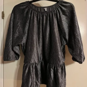 Grå metallic blus med trekvartsärm - Helt ny - Snygg grå metallic blus med rund halsringning och trekvartsärmar. Blusen har ett lätt glansigt material och ett avslappnat fall med resår i midjan för en markerad siluett. Perfekt för dig som vill sticka ut lite extra.