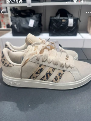 Beige Adidas sneakers med leopardmönster - Snygga beige sneakers från Adidas med klassiska tre ränder i leopardmönster på sidan. Skorna har en låg siluett, matchande beige snörning och detaljer i mockaliknande material. Perfekta för dig som vill sticka ut med en trendig och djurinspirerad look.