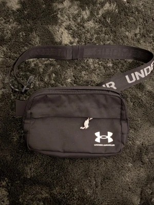 Svart axelväska från Under Armour - Snygg svart axelväska från Under Armour med justerbart band där loggan syns tydligt. Väskan har ett huvudfack med dragkedja och en mindre ficka på baksidan med vit dragkedja. Tillverkad i slitstarkt tyg, perfekt för att bära det viktigaste.
