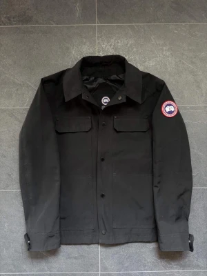 Svart jacka från Canada Goose - Stilren svart jacka från Canada Goose med klassisk krage och två bröstfickor med lock. Jackan har knappstängning framtill och en ikonisk logopatch på vänster ärm. Perfekt för dig som vill ha en enkel men snygg look. Den är för stor för mig så det är därför jag inte använder den längre, använd 3 gånger och kommer inte till nån användning alls 