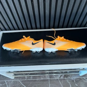 Nike Mercurial orange fotbollsskor (helt oanvända) - Säljer ett par grymma Nike Mercurial fotbollsskor i orange med vita och svarta detaljer. Skorna har klassisk låg profil, snörning och dobbar för spel på gräs. Tillverkade i syntetmaterial för lätt känsla och bra bollkontroll. Perfekta för dig som vill sticka ut på planen.