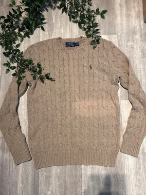 Beige kabelstickad tröja Polo Ralph Lauren - Kabelstickad beige tröja från Polo Ralph Lauren med rund halsringning och diskret broderad logga på bröstet. Tröjan har långa ärmar och ribbade muddar vid ärmslut och nederkant. Perfekt för en stilren och klassisk look.