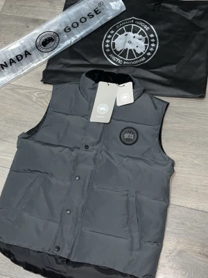Svart dunväst från Canada Goose - Snygg svart dunväst från Canada Goose med diskret logga på bröstet. Västen har två sidofickor, tryckknappar och dragkedja framtill. Tillverkad i slitstarkt material och fylld med dun för extra värme. Perfekt för lager-på-lager och streetwear-stil.