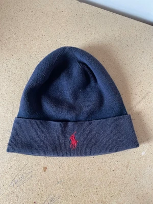 Mörkblå mössa från Polo Ralph Lauren - Snygg mörkblå mössa från Polo Ralph Lauren med klassisk uppvikt kant och röd broderad logga framtill. Tillverkad i mjukt stickat material som håller dig varm och stilren under kalla dagar. Perfekt accessoar för dig som gillar enkel och tidlös design.