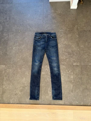 Mörkblå skinny jeans från Nudie - Säljer ett par mörkblå skinny jeans från Nudie Jeans med klassisk femficksmodell och slitna detaljer. Jeansen har en snygg tvätt med ljusare partier och är tillverkade i  denim. Modellen är grimtim och är i helt nytt skick förutom fadsen byxorna har fått övertid. Sitter dom 30/30 i storleken