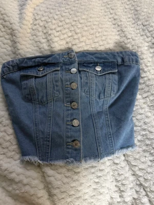 Blå jeans tubtopp - Säljer en snygg blå jeans tubtopp med knappar framtill, två bröstfickor och rå kant nertill. Toppen är axelbandslös och har smock baktill för skön passform. Perfekt för dig som vill ha en trendig och edgy look.