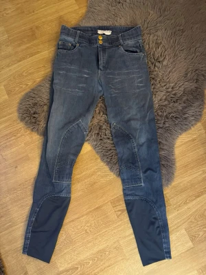 Mörkblå ridbyxor  - Säljer ett par mörkblå ridbyxor med förstärkta partier på knän och insida ben. Byxorna har två guldknappar i midjan, klassiska fickor fram och kontrastmaterial nedtill på benen. Snygga detaljer med slitningar framtill och snygga fickor baktill.