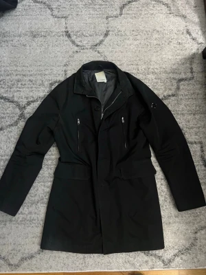 Svart rock från J.Lindeberg - Stilren svart rock från J.Lindeberg med två snedställda bröstfickor med dragkedja och klassisk krage. Rocken har en rak passform och är fodrad på insidan för extra komfort. Perfekt för dig som vill ha en modern och enkel look.