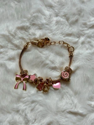 Guldigt charmarmband med rosa detaljer - Snyggt guldigt armband med flera rosa berlocker i form av rosett, stjärna, blomma, hjärta och klubba. Armbandet har glittriga pärldetaljer och justerbar kedja. Perfekt accessoar för dig som gillar söta och lekfulla smycken.