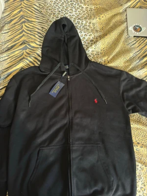 Svart Polo Ralph Lauren - Säljer en svart hoodie från Polo Ralph Lauren med dragkedja framtill och klassisk röd logga broderad på bröstet. HELT NY OANVÄND, KOM MED BUD
