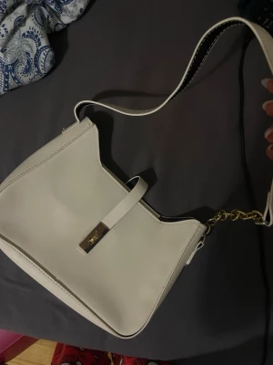 Vit axelväska med gulddetaljer - Snygg vit handväska med stilren design och guldfärgad kedja på axelremmen. Väskan har ett dekorativt spänne framtill och är tillverkad i slätt konstläder. Perfekt för dig som gillar minimalistisk och trendig stil.
