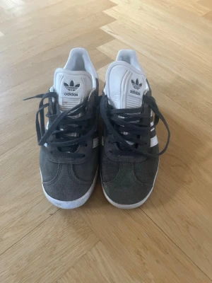 Adidas Gazelle grå sneakers - Snygga Adidas Gazelle sneakers i grå mocka med vita detaljer och klassiska tre ränder på sidan. Skorna har svart snörning, vit sula och Adidas-logga på plösen. Använda ett par gånger, säljer pga för små 
