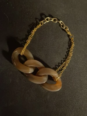 Armband med stora länkar i brunt - Snyggt armband med tre stora, bruna länkar i plast och dubbla guldfärgade kedjor. Länkarna har en mjuk, rundad form och armbandet stängs med en karbinhake. Perfekt accessoar för att lyfta din outfit.